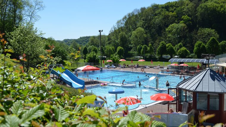 Ein Schwimmbad mit einer Rutsche und vielen Sonnenschirmen. Menschen genießen das Wasser und die Sonne in einer grünen Umgebung.