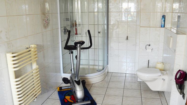 A modern bathroom with a shower cubicle, a toilet area, and a fitness machine. The room is bright and has large tiles.