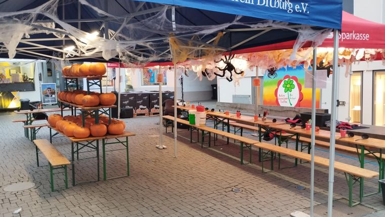 Ein Veranstaltungsbereich mit mehreren Tischen und Bänken. In der Mitte stehen orange Kürbisse und dekorative Spinnweben sind sichtbar.