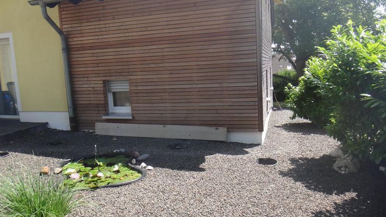 Un espace extérieur calme avec un revêtement en bois et une surface en gravier. Sur le côté gauche, un petit étang avec des plantes est visible.
