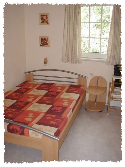 Une chambre simple avec un lit et une petite table. Les murs sont clairs et il y a des fenêtres avec des rideaux.