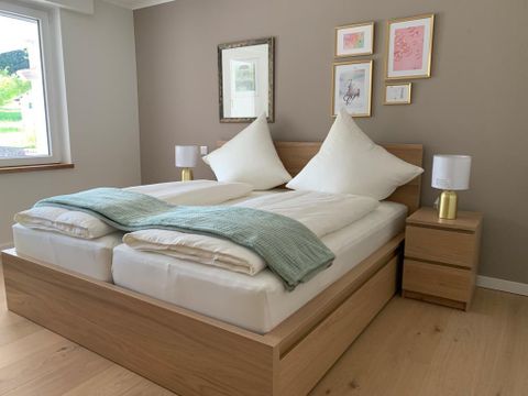 Ein modernes Schlafzimmer mit einem Doppelbett und großen, gemütlichen Kissen. An der Wand hängen gerahmte Bilder und die Fenster bieten einen Blick nach draußen.