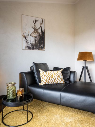 Un salon moderne avec un canapé noir et un coussin coloré.
Au mur, il y a une image d'un cerf et il y a une lampe sur pied ainsi qu'une petite table.