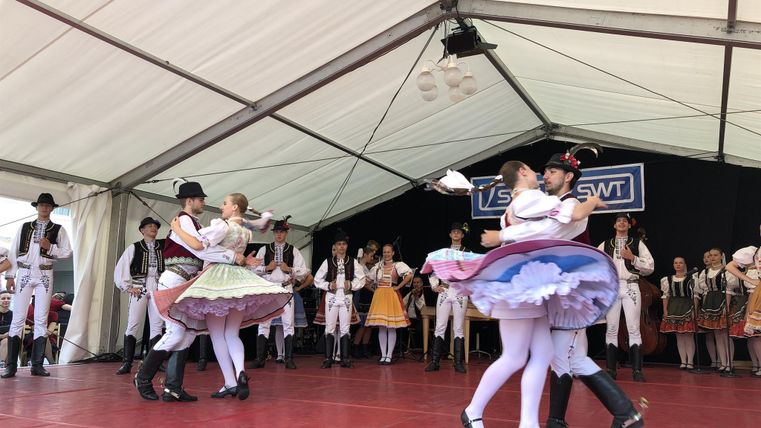Eine Gruppe von Tänzern in traditionellen Kostümen führt einen Volkstanz auf einer Bühne auf. Im Hintergrund sind weitere Tänzer und Musiker zu sehen.