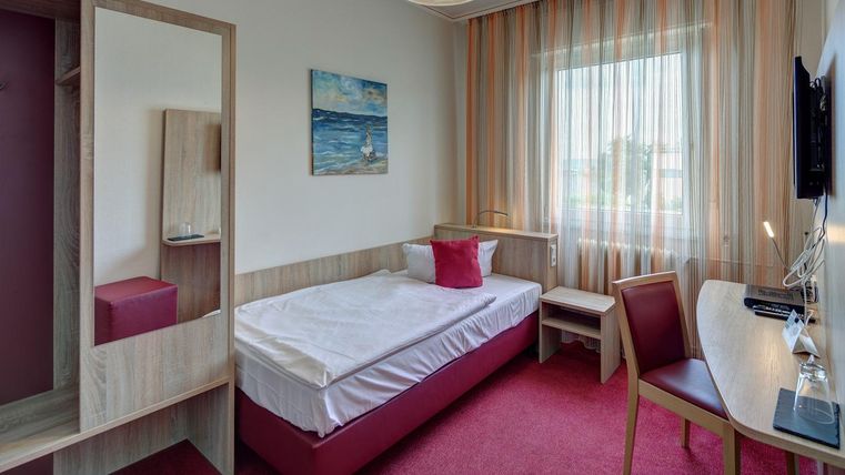 Une chambre d'hôtel moderne avec un lit simple et un petit bureau. La grande baie vitrée laisse entrer beaucoup de lumière naturelle et crée une atmosphère agréable.