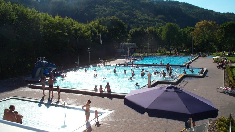 Een zwembad met meerdere bassins en veel mensen die daar zwemmen en spelen. Op de achtergrond zijn groene heuvels en bomen te zien.