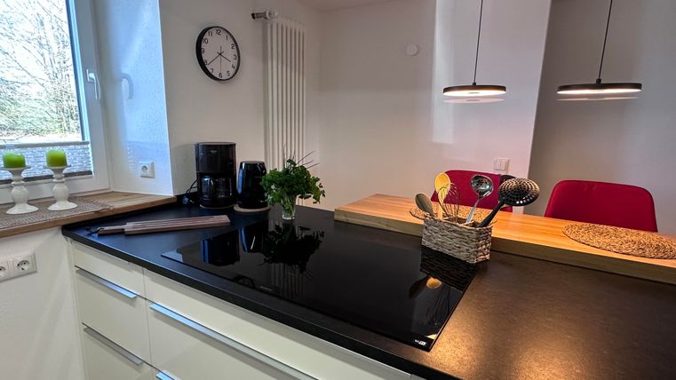 Eine moderne Küche mit einer schwarzen Arbeitsplatte und minimalistischen weißen Schränken. Im Hintergrund sind Fenster und dekorative Elemente zu sehen.