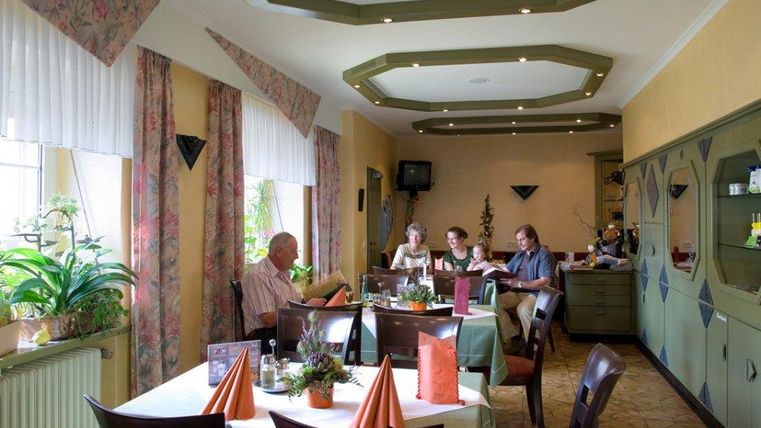 Ein gemütliches Restaurant mit vier Gästen, die an einem Tisch sitzen. Die Einrichtung ist freundlich, mit Pflanzen und farbigen Tischdecken.