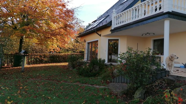 Ein gemütliches Haus mit einem Balkon, umgeben von herbstlichen Bäumen. Der Garten ist grün und es gibt einen Hund auf der Terrasse.