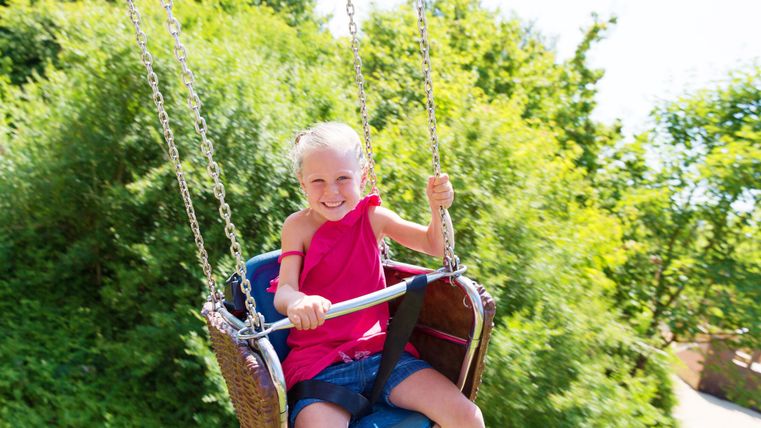 Ein fröhliches Kind schwingt auf einer Schaukel im Freien. Die Umgebung ist grün und sonnig.