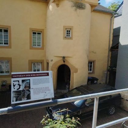 Ein historisches Gebäude mit gelber Fassade und einem Eingang. Im Vordergrund steht ein Informationsschild.