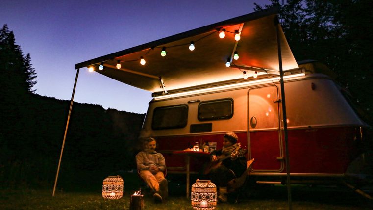 Een gezellige campingavond onder een tent met lichten. Twee personen zitten ontspannen naast een caravan in de natuur.