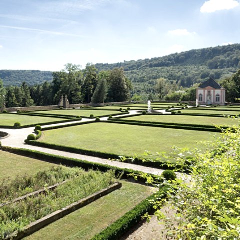 French garden with symmetrical hedges and lawns, a small building in the background and wooded hills under a blue sky., &copy; V. Teuschler