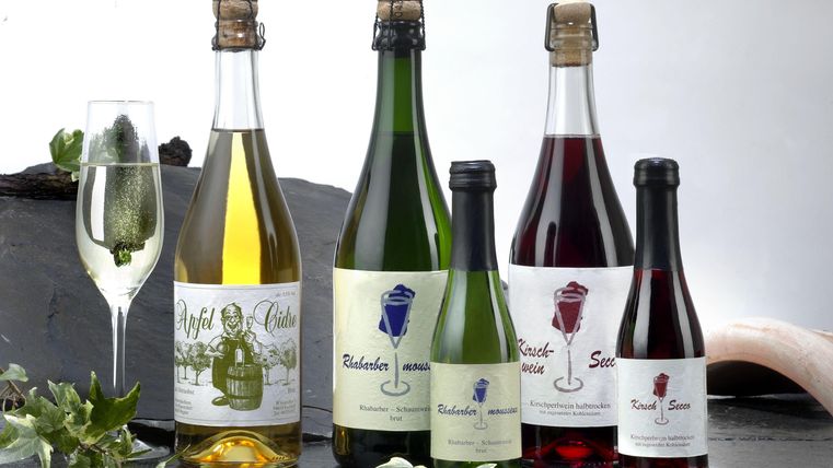 A selection of bottles with various drinks, including non-alcoholic juices and sparkling wines. In the foreground is a glass with a drink and a slice of cucumber.