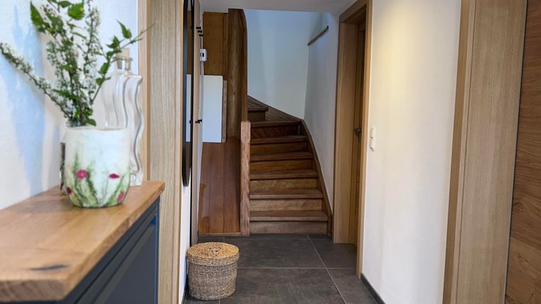 Ein heller Flur mit einer Holztreppe und modernen Wänden. Auf einem Tisch steht eine Blumenvase und ein Korb liegt auf dem Boden.