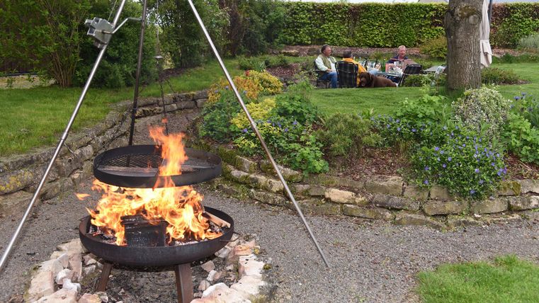 Un jardin accueillant avec un feu ouvert dans un barbecue. À l'arrière-plan, des personnes sont assises confortablement sur une pelouse.
