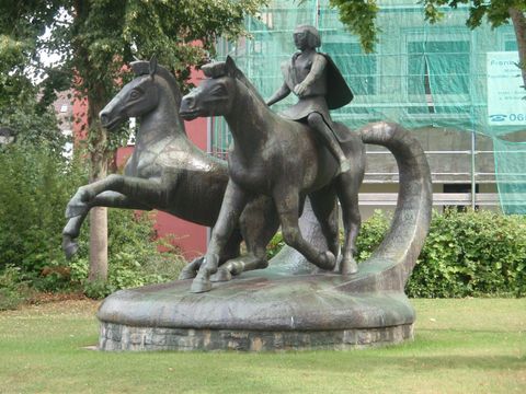 A bronze statue shows a rider on two horses. The statue stands on a green lawn outdoors.