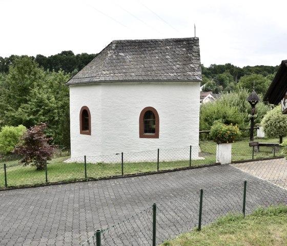 White chapel with slate roof, surrounded by green landscape. A sign with the inscription "Kapellenhof" stands in the foreground., © TI Bitburger Land