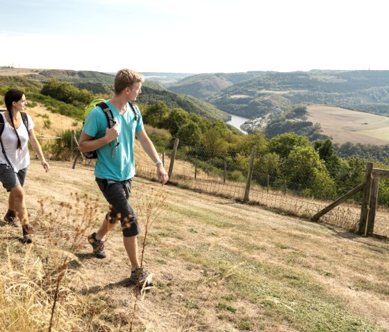 Zwei Personen wandern auf einem Hügel mit Blick auf eine Flusslandschaft. Die Umgebung ist grün und hügelig, der Himmel ist klar., © Eifel Tourismus GmbH, D. Ketz