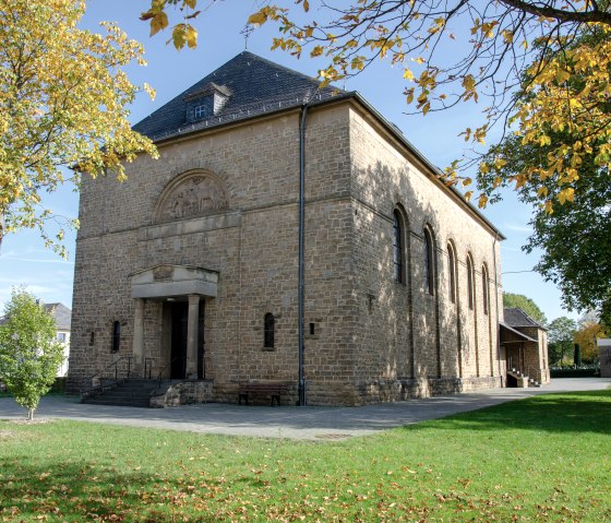 The church of St. Hubertus in Wolsfeld, surrounded by autumnal trees, stands in a green park. The building is made of stone with a gabled roof., © TI Bitburger Land