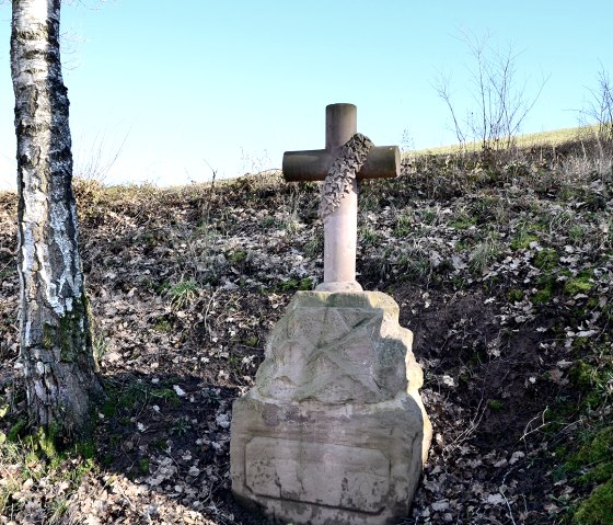 Ein steinernes Wegekreuz mit Efeu bewachsen steht zwischen zwei Bäumen auf einem bewachsenen Hügel., © TI Bitburger Land