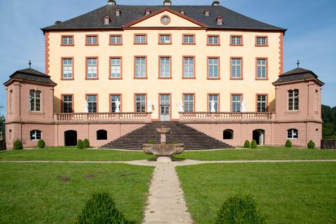 Ein elegantes Schloss mit einer hellen Fassade und großen Fenstern. Der gepflegte Garten mit einem Weg und einem Brunnen lädt zum Verweilen ein.