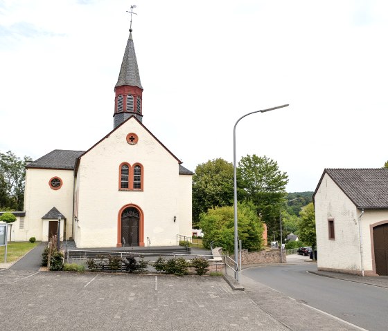 Eine Kirche in Wi&szlig;mannsdorf mit spitzem Turm, umgeben von B&auml;umen. Davor ein leerer Parkplatz und eine Stra&szlig;e. Rechts ein weiteres Geb&auml;ude., &copy; TI Bitburger Land