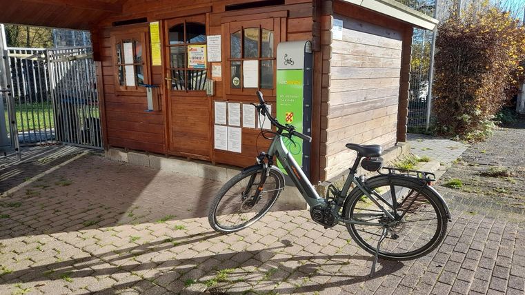 Een elektrische fiets staat voor een houten gebouw met ramen en een informatiebord. Het weer is zonnig en de grond is bedekt met wat bladeren.