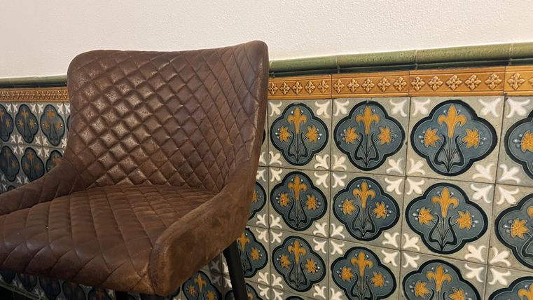 Een stijlvolle stoel met een gepolsterde bovenkant staat voor een kleurrijke tegelwand. De tegels tonen een aantrekkelijk patroon in blauw en geel.
