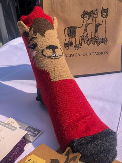 Une chaussette colorée avec un motif de lama en rouge et marron. En arrière-plan, d'autres produits et un sac avec l'inscription "ALPACA. OUR PASSION." sont visibles.