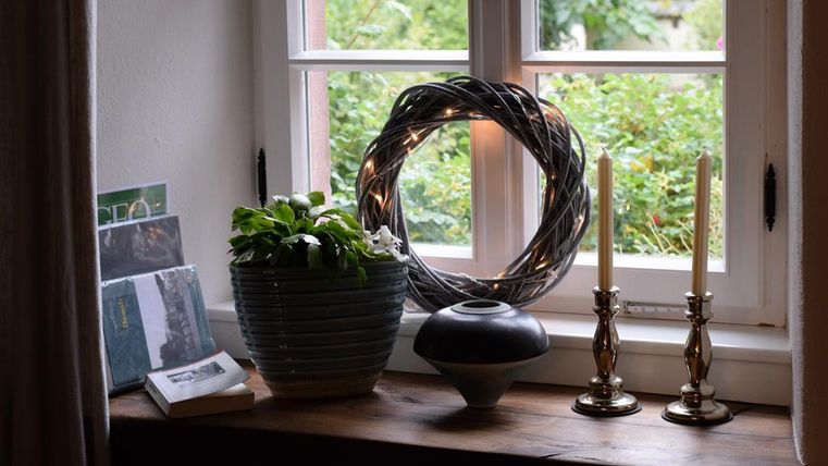 Eine Fensterbank mit einer blühenden Pflanze in einem Topf und Kerzenhaltern. Im Hintergrund ist ein dekorativer Kranz und einige Karten zu sehen.