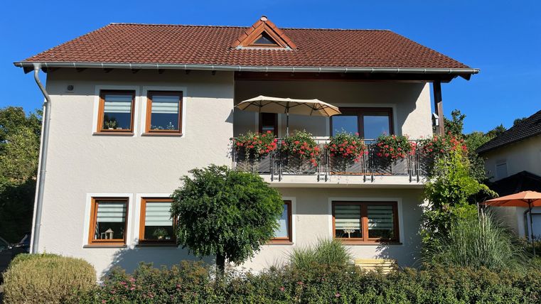 Une jolie maison avec un toit rouge et des jardinières adjacentes. Le jardin est vert et soigné, avec des buissons et un petit arbre.