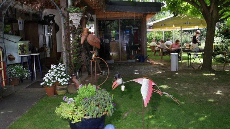 Un jardin accueillant avec des plantes colorées et une fontaine qui coule. À l'arrière-plan, on peut voir des places assises sous un parasol et une atmosphère conviviale.