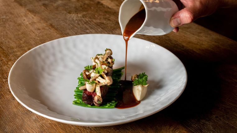 Een kunstzinnig opgemaakt bord met groenten en saus. Een hand giet de saus over het gerecht.