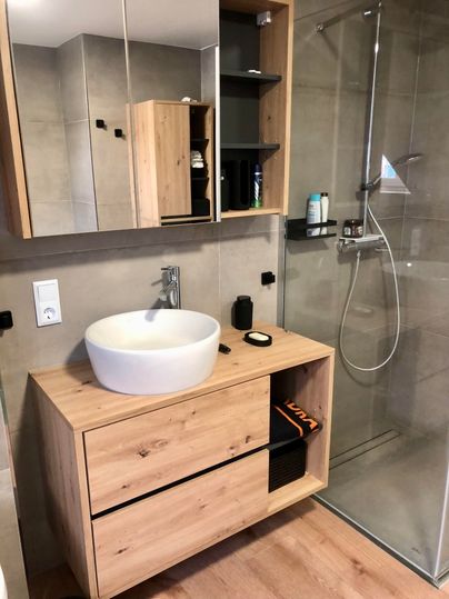 A modern bathroom with a wooden sink cabinet and a round washbasin. There is a glass shower and practical shelves for storage.