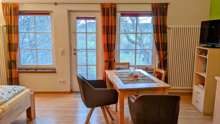 Ein gemütlicher Wohnraum mit einem Tisch und Stühlen. Die Fenster bieten einen Blick ins Freie und sind mit bunten Vorhängen dekoriert.