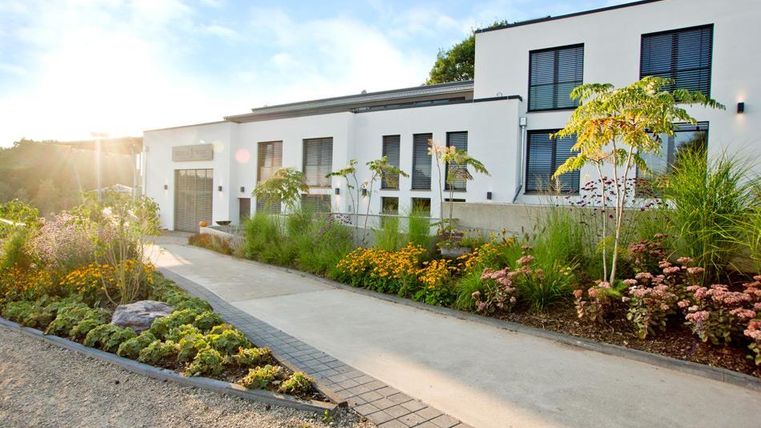 Un bâtiment moderne avec de grandes fenêtres et une entrée accueillante. Le jardin est luxuriant avec des fleurs colorées et des plantes vertes.