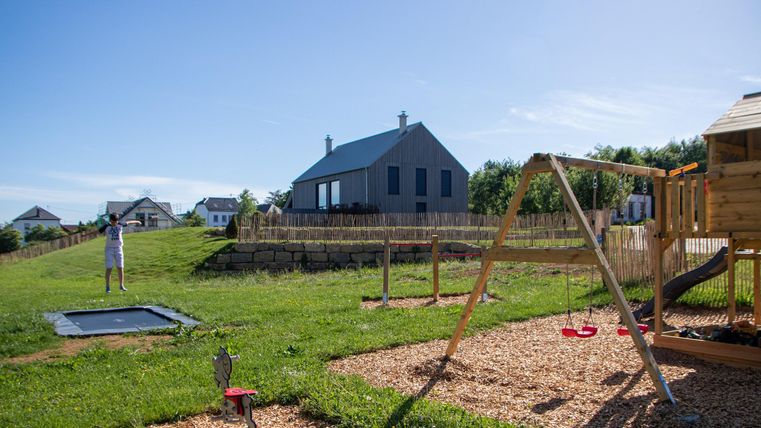 Ein schöner Spielplatz mit einer Schaukel, einem Trampolin und Spielgeräten aus Holz. Im Hintergrund steht ein modernes Haus und der Himmel ist klar und blau.