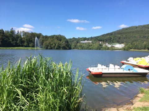 Un lac paisible entouré de rives verdoyantes et de douces collines. On peut voir quelques pédalos sur l'eau, et le ciel est clair et bleu.