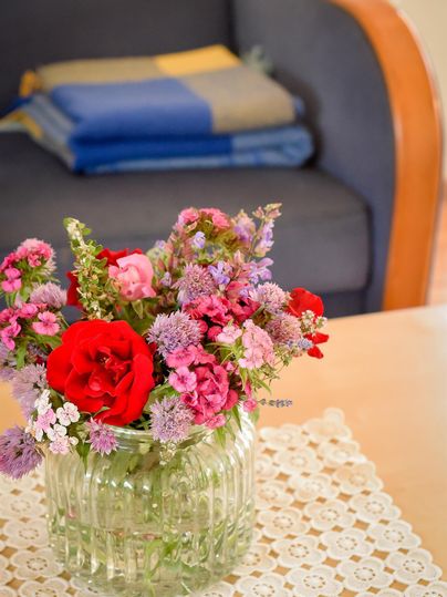 Een mooie bloemenboeket met rode en roze bloemen in een heldere glazen vaas. Op de achtergrond zijn een sofa en een opgevouwen deken te zien.