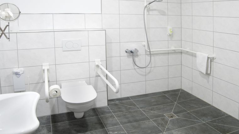 Une salle de bain moderne accessible aux personnes à mobilité réduite avec des carreaux blancs et un sol gris. Elle comprend des toilettes, une douche et des barres d'appui pour plus de sécurité.