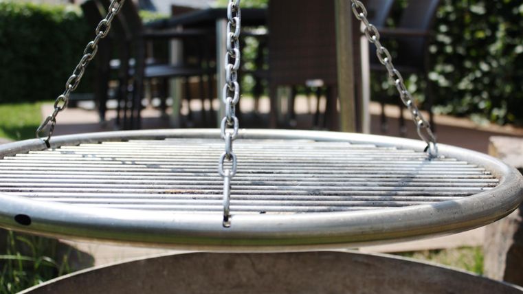 Ein runder Grill mit Grillrosten hängt an Ketten. Im Hintergrund sind Gartenmöbel zu sehen.