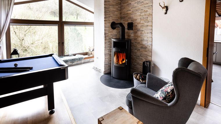 A cozy living room with a fireplace and a billiard table. Large windows offer a view of the nature outside.