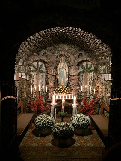 Ein geschmücktes Heiligtum mit einer Statue einer Heiligen, umgeben von Kerzen und Blumen. Die Wand ist mit Steinen verziert und es gibt zwei große Pflanzen auf beiden Seiten.