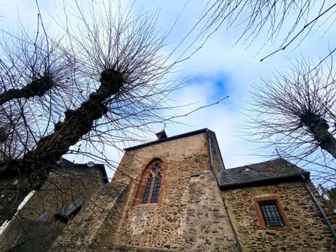 Eine alte Kirche mit einer steinernen Fassade und einem schönen Fenster. Im Vordergrund sind kahle Bäume sichtbar und der Himmel ist bewölkt.