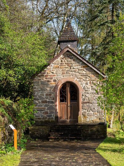 Eine kleine Stein-Kapelle umgeben von grünem Laub und Bäumen. Der Eingang hat eine elegante Holztür und die Architektur ist schlicht und charmant.