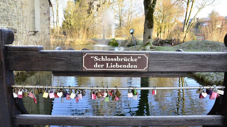 Eine romantische Brücke mit einem Schild, das "Schlossbrücke der Liebenden" zeigt. An der Brücke hängen viele Vorhängeschlösser, die Liebe symbolisieren.