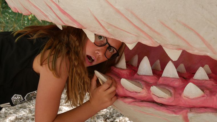 Een meisje met een bril poseert in een kunstzinnige model van een haaienmond. Ze kijkt verrast uit de bek van de haai.