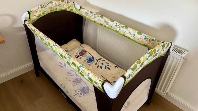 Ein Babybett mit einer bunten Liegematte und weichen Rändern steht in einem hellen Raum. Der Boden ist aus Holz, und das Bett ist gut beleuchtet.