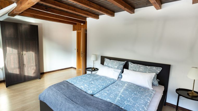 A modern bedroom with a large double bed and wooden beam ceiling. There is a black wardrobe against the wall and two bedside tables with lamps.